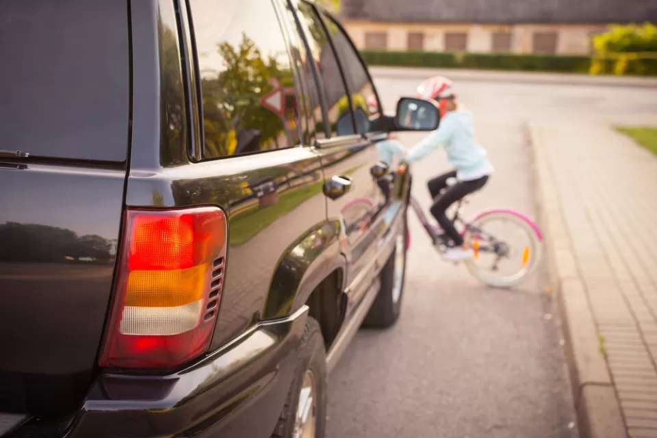 View of large black SUV from behind with blurred small child on bicycle wearing helmet riding in front of car